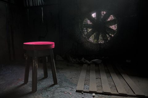 Light flows in through a large fan covered with mesh, it illuminates the red top of a stool, the only bit of colour in the picture.