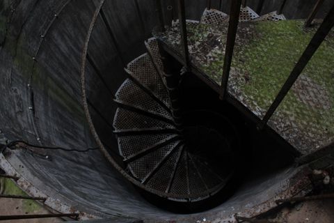 A metal staircase wound in a tight spiral descends into a circular structure below, no bottom is visible, the steps simply disappear into blackness.