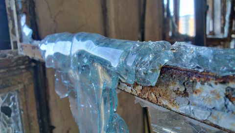 A close up of smooth, translucent blue glass that has half melted and appears to ooze over the edge of its rusty, fire damaged frame.