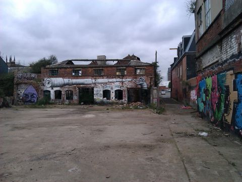 An empty yard leads to a derelict factory building missing most of its roof. A painting by street artist Phlegm features a hooded figure holding a telescope which spans the width of the building.