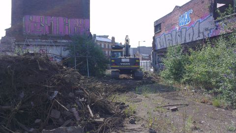 The main building is visible with new scaffold supports. In front of it is a mound of rubble and plants which have been cleared away. The other building is visible on the right side, half hidden by buddleia. A digger sits between the two.