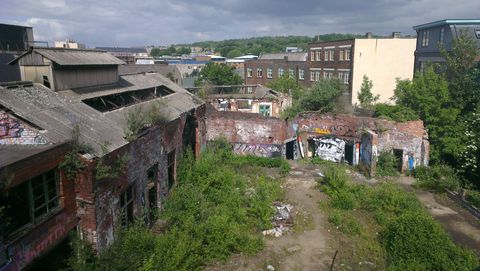 An elevated view of the whole site from within. On the left, a three-storey building with a swathe of missing brickwork forming a huge arch. Straight ahead, the building I started in joins it to form an L shape. Green double doors are just visible on the upper floor.