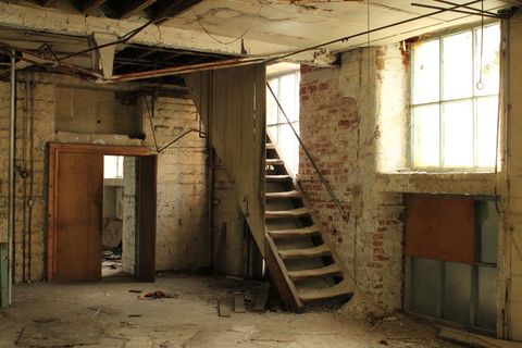 Soft, yellow light filters in through windows, illuminating an empty room in a mill. A staircase disappears upwards, each step is worn in the middle from countless steps and is piled high with dust.
