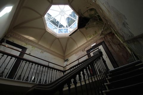 Light streams in through an octagonal skylight set into a vaulted ceiling, one quarter of which is green and water damaged with a gaping hole, while the rest still has almost pristine ornamental cornicing. Twisted wrought iron banisters wrap the landing and descend the stairs.