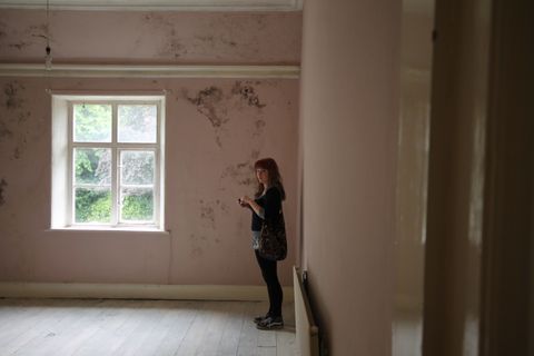 A girl stands in a bare bedroom at the centre of the shot glancing sidelong at the camera, framed by faded pink walls that are beginning to mould. Soft light filters in through a large window, offering a glimpse of dense shrubbery in the garden beyond.