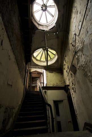 A vertical, wide-angle shot beginning at the bottom of a narrow staircase, ascending to an open door set in large frame with a triangular lintel. Two large, round skylights above illuminate the scene, with algae growth tinting the flaking walls a yellowy-green.