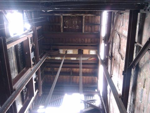 Looking up to the top of a lift shift, the pulleys and beams are rusted with age. Sunlight streams in through gaps in the corrugated sheeting covering the tops of the walls.
