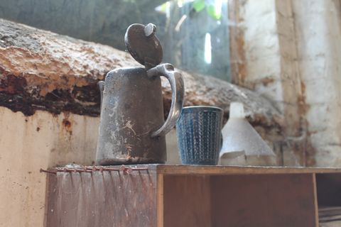 A tarnished metal teapot and accompanying blue mug sit on a cupboard, there are rusty nails all along the side of it for hanging tools or tea towels.