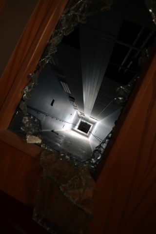 This shot is twisted sideways, showing a long corridor extending through the smashed reinforced glass of a fire door. A broken section of ceiling lights a small band of the corridor dramatically before it plunges into darkness at the end.