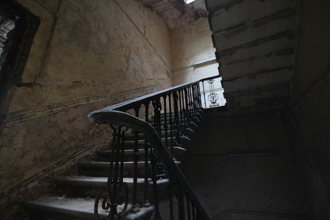 A stone staircase with ornate iron bannisters, the walls are yellowed and cracked where panelling used to be.